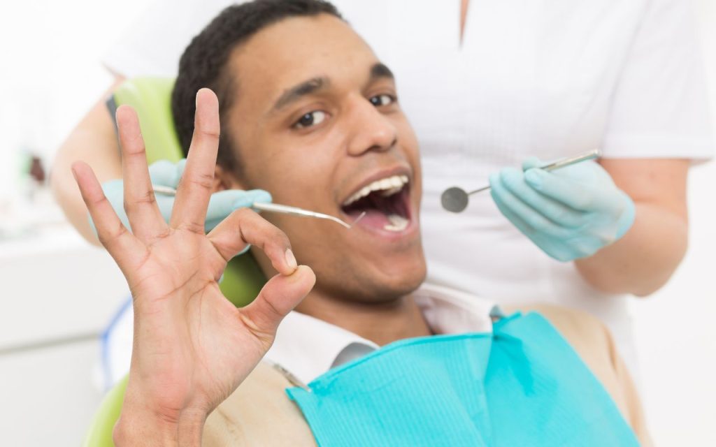 Preventative dental care - A man sitting in a dental chair giving an "OK" hand gesture while a dentist behind him holds dental instruments.