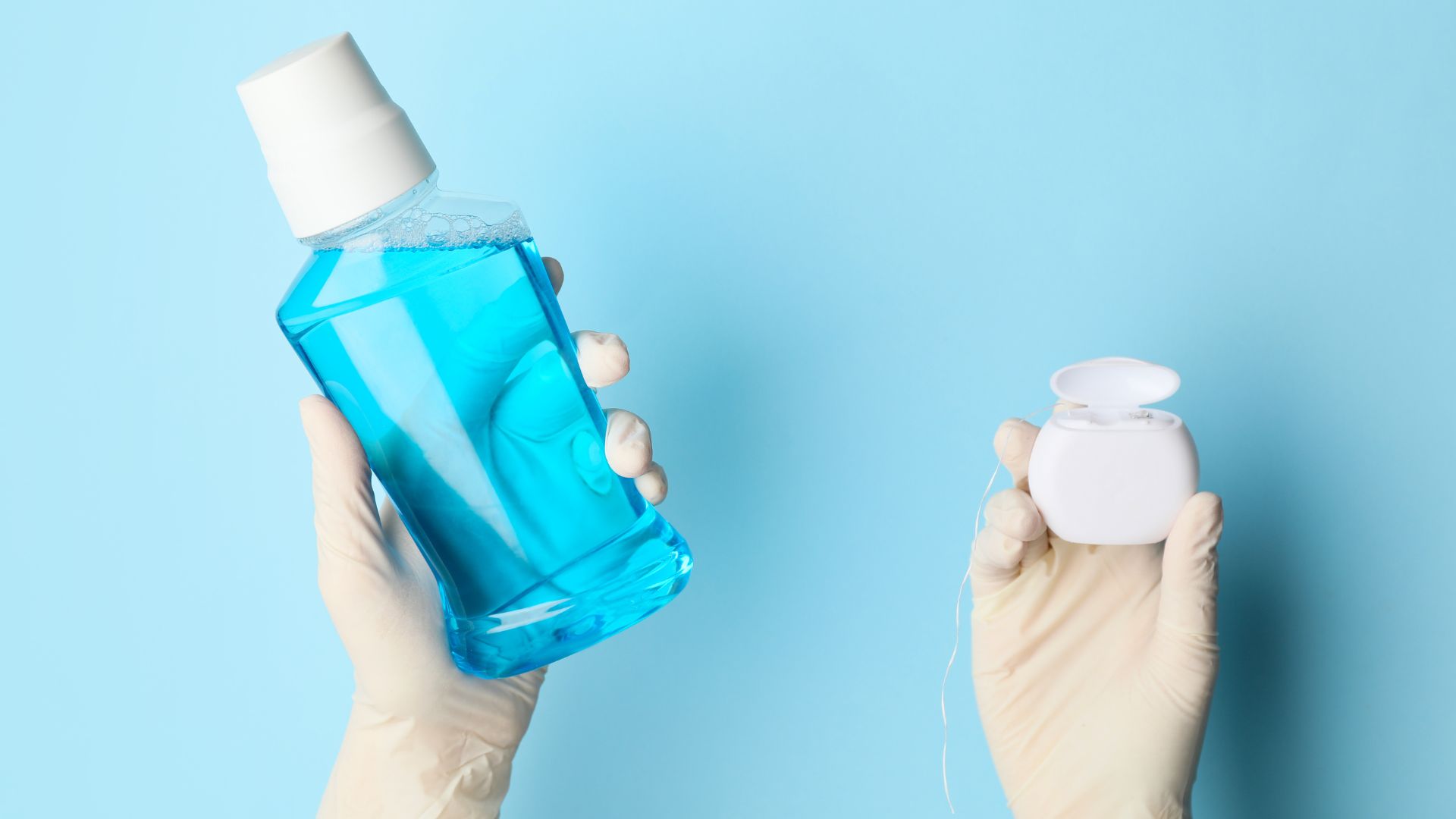 Gloved hands holding a bottle of blue mouthwash in one hand and dental floss in a white container in the other, against a light blue background.