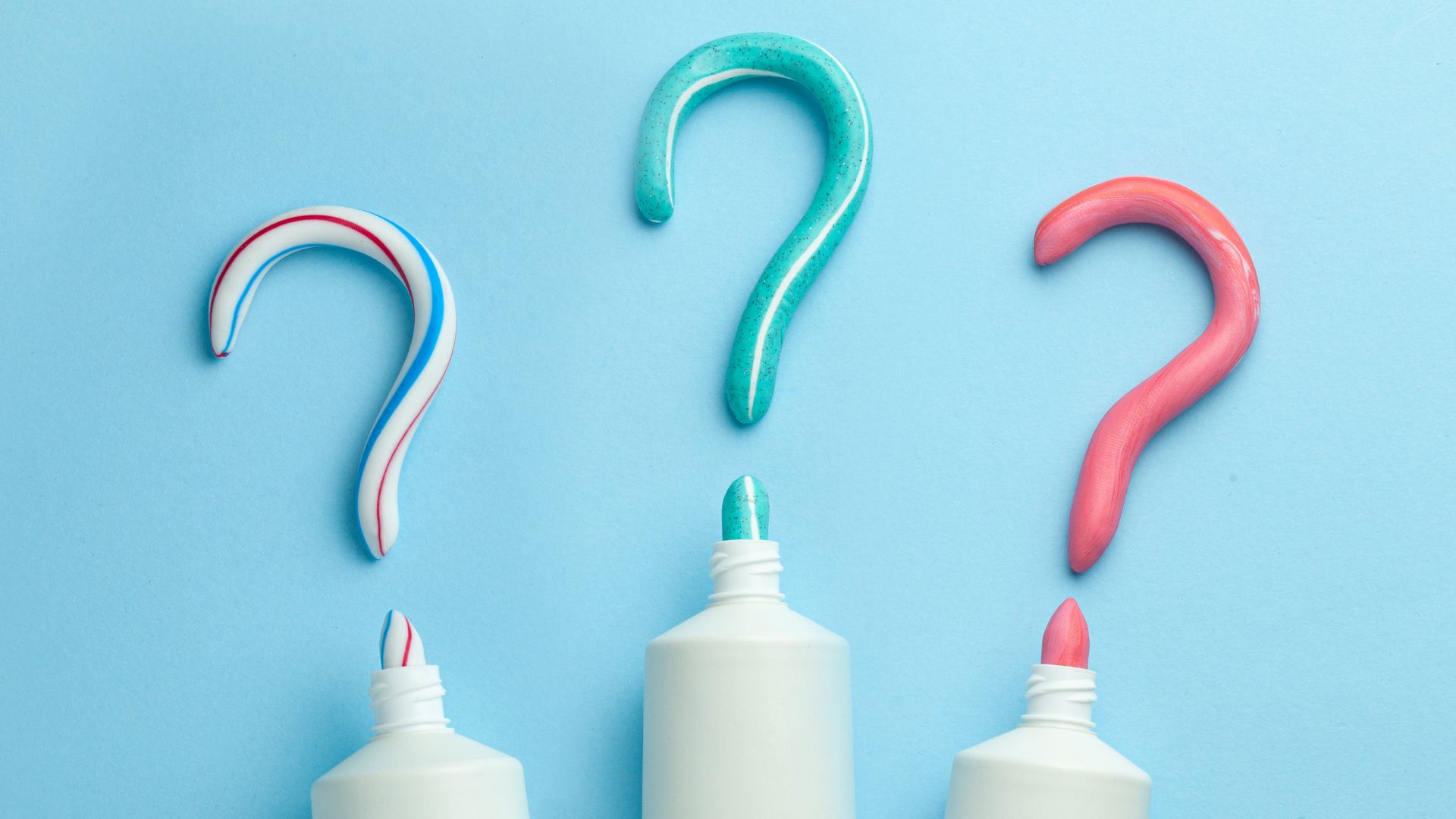 Three toothpaste tubes squeeze out toothpaste shaped as question marks in different colors—red, green, and red-white-blue—on a light blue background.