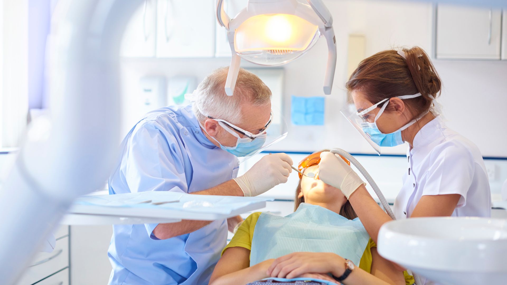 Two dentists wearing masks and gloves perform a dental procedure on a patient lying in a dental chair under a bright examination light.