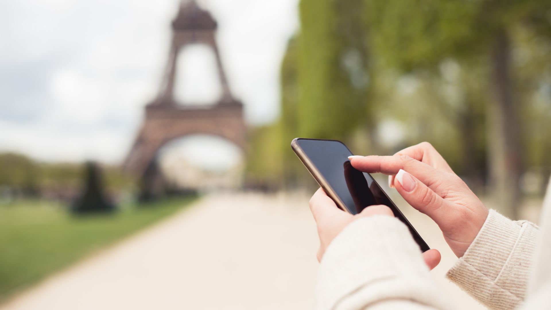 A person holds and uses a smartphone outdoors with the blurred Eiffel Tower visible in the background, possibly searching how to find a dentist abroad during a dental emergency while traveling.