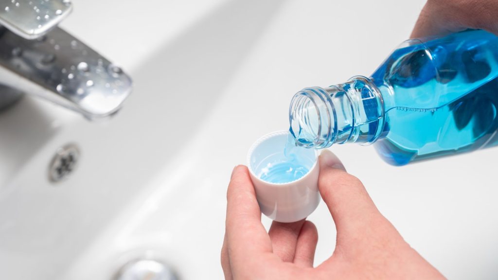 A person pours blue mouthwash from a bottle into the cap over a white sink.
