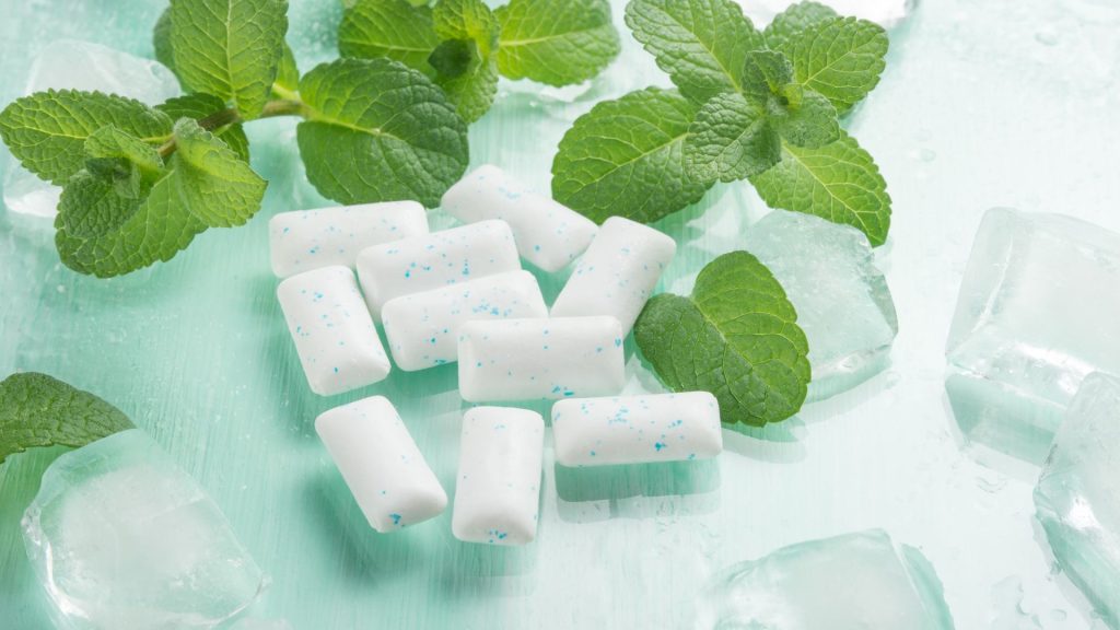 Rectangular pieces of white chewing gum with blue specks are surrounded by mint leaves and ice cubes on a light green surface.