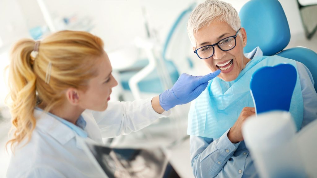 A dentist examines an older patient's teeth while the patient holds a mirror, both seated in a dental clinic.