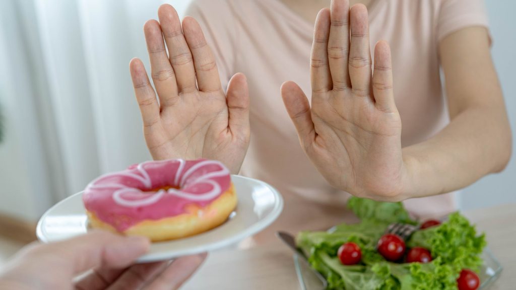 What an ideal healthy oral routine looks like: A person raises both hands to refuse a pink frosted donut on a plate, with a salad in front of them on the table.