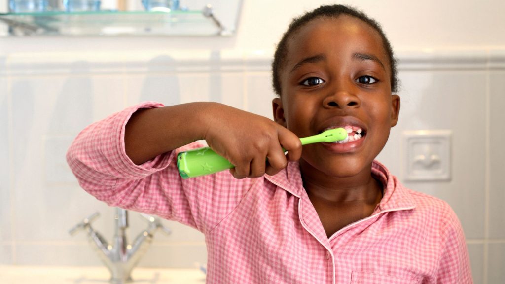 A child in a pink shirt is brushing their teeth with a green toothbrush in a bathroom.