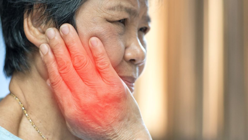 An older woman holds her hand to her cheek, which is highlighted red, suggesting pain or discomfort in her jaw or face.