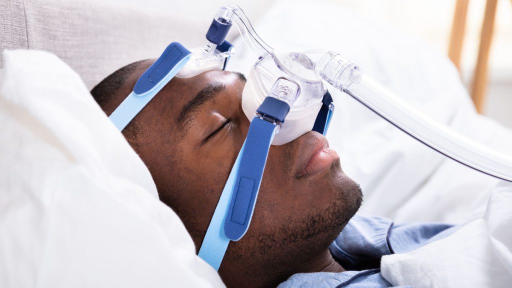A person is lying in bed wearing a CPAP mask connected to tubes, commonly used for sleep apnea treatment.