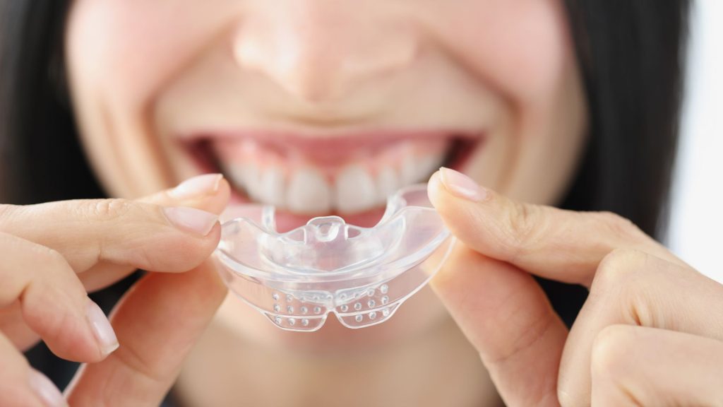 A person holds a clear dental mouthguard near their smiling mouth, preparing to insert it over their teeth.