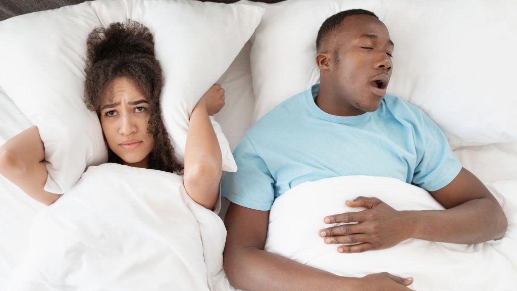 A woman covers her ears with a pillow while lying in bed next to a man who is sleeping with his mouth open, possibly snoring.