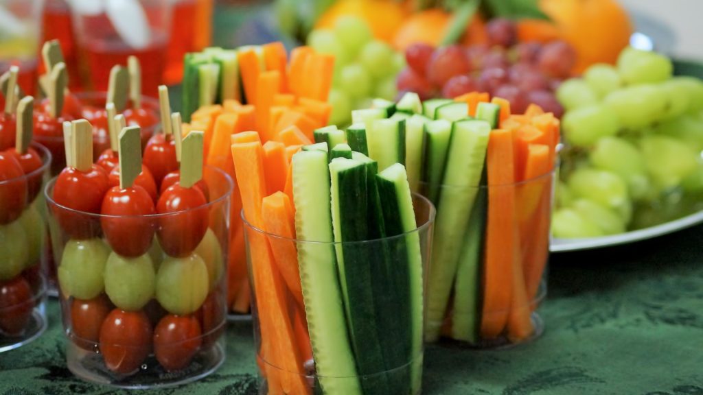 Clear plastic cups filled with grape tomatoes, green grapes, carrot sticks, and cucumber sticks are arranged on a table with more fruit in the background.