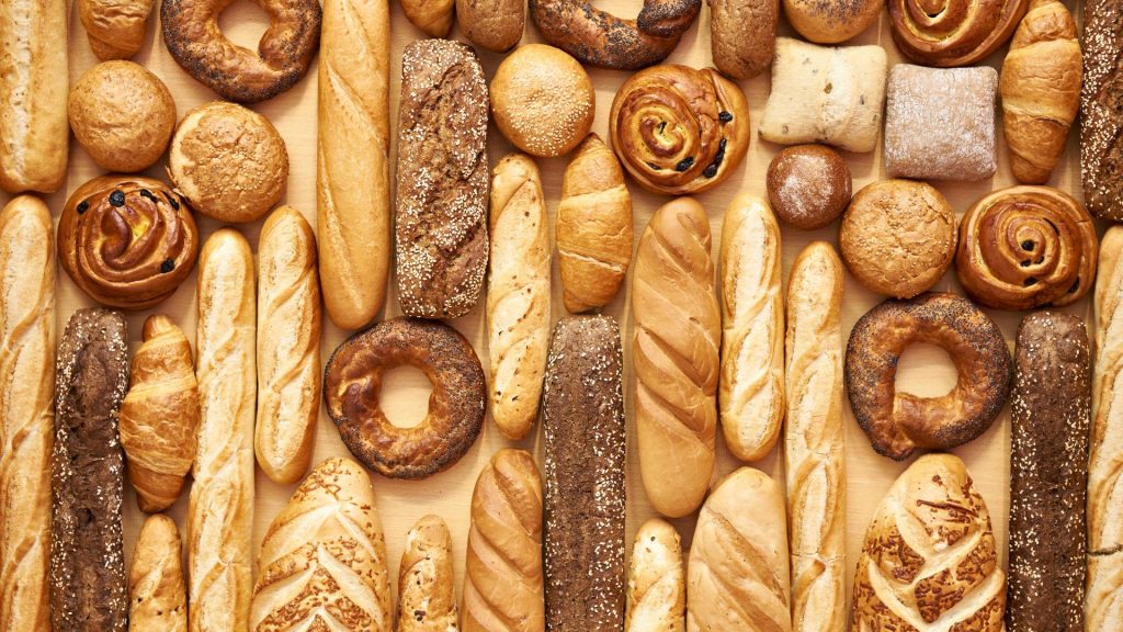 An assortment of various breads and pastries, including baguettes, rolls, bagels, and loaves—some known as foods that can crack your teeth—arranged in rows on a wooden surface.
