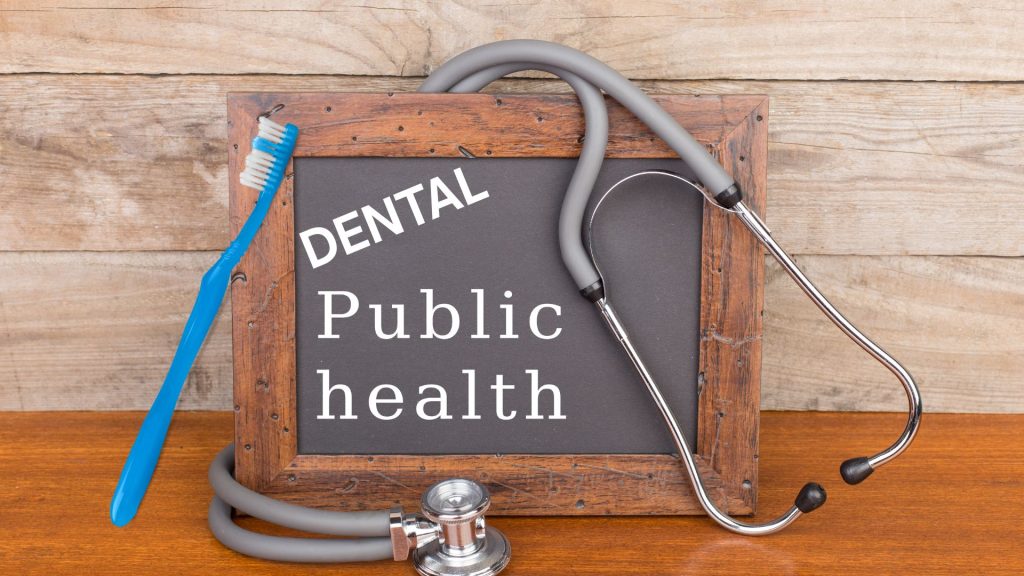 A chalkboard with the words "Dental Public Health" is displayed alongside a stethoscope and a toothbrush on a wooden surface.