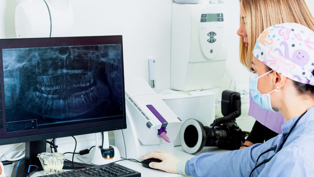 Two dental professionals examine a dental X-ray displayed on a computer monitor in a clinical setting.