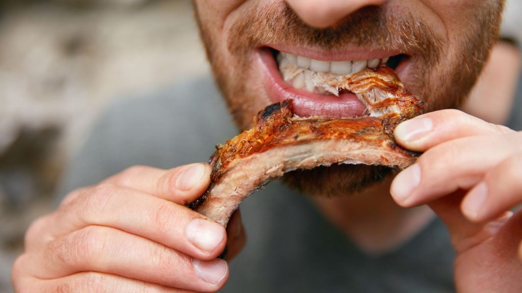 A person eating grilled meat from a rib bone, close-up of mouth and hands—reminding us how hard foods that damage teeth, like ribs, can sometimes lead to a cracked tooth.