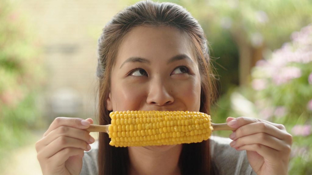 A person holding and eating a corn on the cob, looking upwards, with a blurred outdoor background—a reminder that certain foods that can crack your teeth require extra care when biting in.