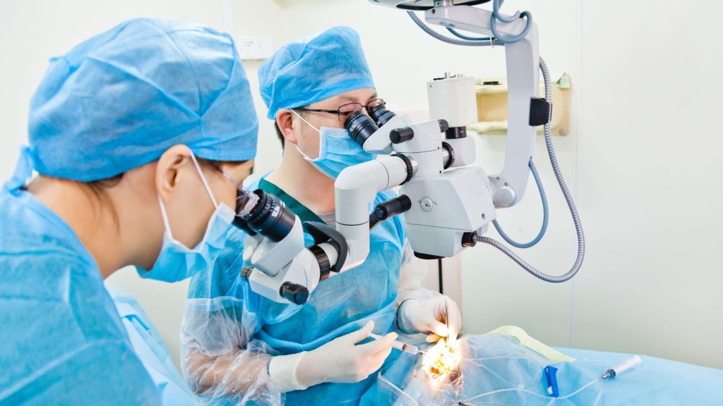 Two surgeons in blue scrubs and masks use a microscope and surgical tools during a procedure in an operating room.