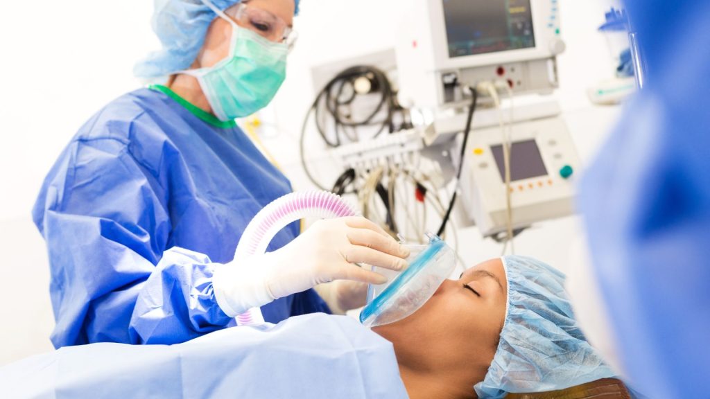 Medical professional in blue scrubs and a mask administering anesthesia to a patient lying on an operating table in a hospital setting with medical equipment in the background.