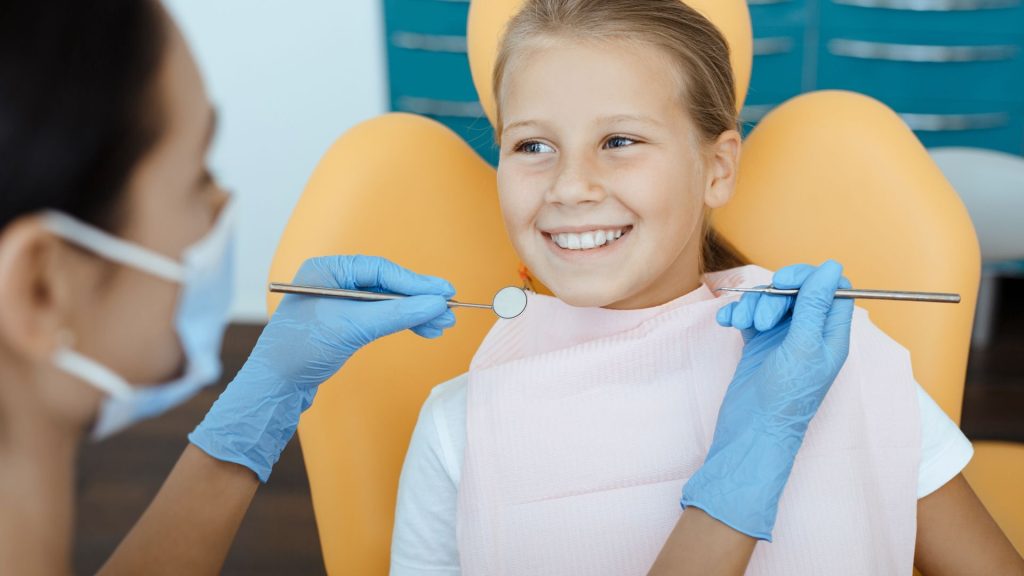 A dentist wearing gloves examines a smiling girl’s teeth with dental instruments while she sits in an orange dental chair.
