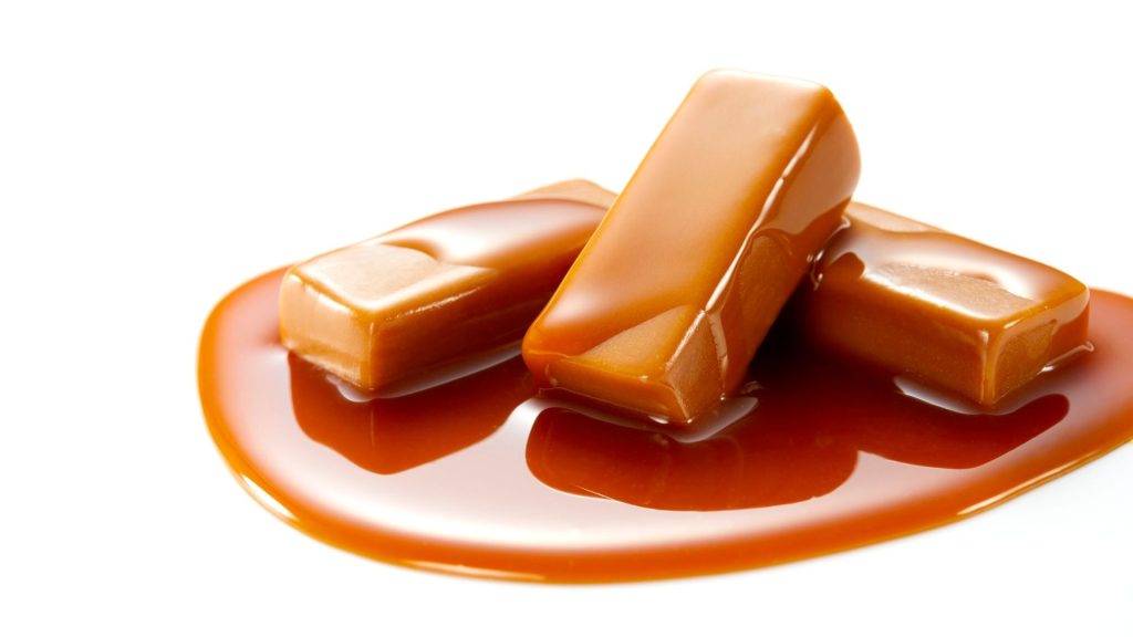 Three rectangular caramel candies sit on a pool of melted caramel sauce against a white background, reminding us that sticky treats can be among the foods that can crack your teeth or even cause tooth fractures if eaten too often.