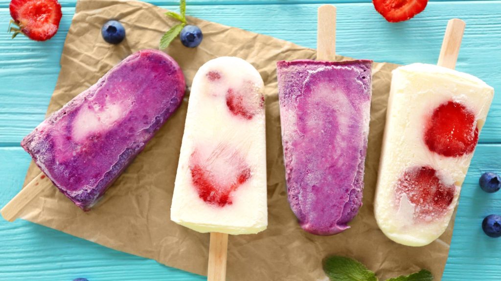 Four fruit popsicles with swirled colors and pieces of berries are arranged on brown paper, surrounded by scattered blueberries and mint leaves on a blue wooden surface—reminding us that some frozen treats can be among the foods that crack teeth.