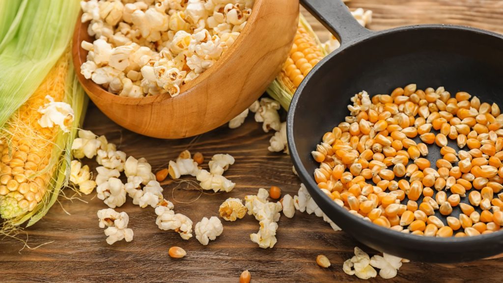 A wooden bowl of popped popcorn, a black pan filled with unpopped corn kernels, and corn cobs on a wooden surface highlight foods that can crack your teeth if you’re not careful.