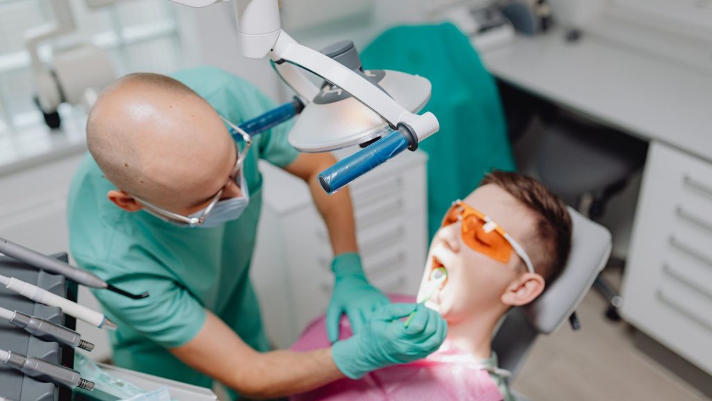 A dentist wearing gloves and a mask examines a patient's mouth for signs of a cracked tooth in a dental clinic, with the patient wearing protective glasses.