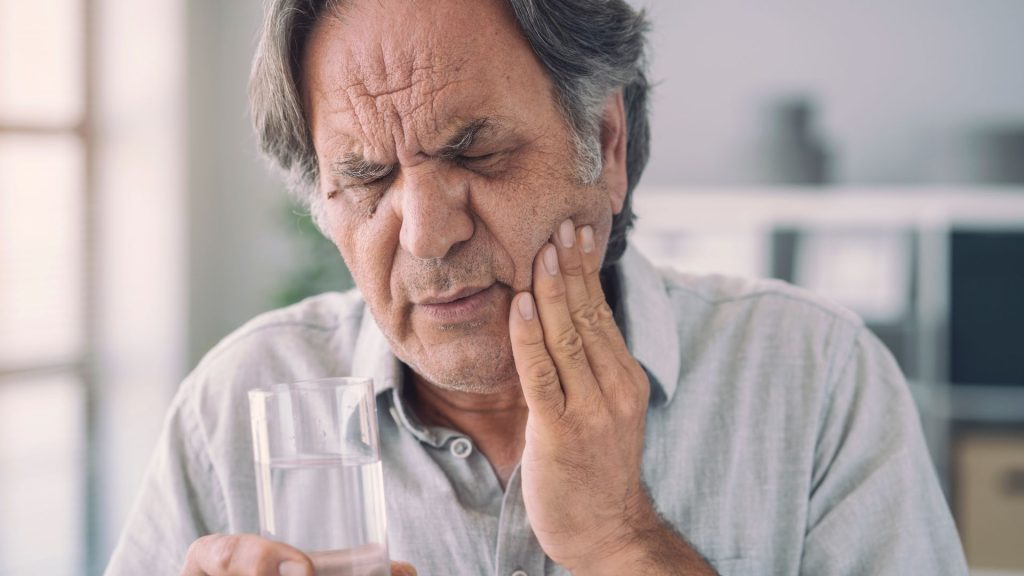 An older man holds a glass of water in one hand and touches his cheek with a pained expression, suggesting discomfort from a cracked tooth or other dental issues.