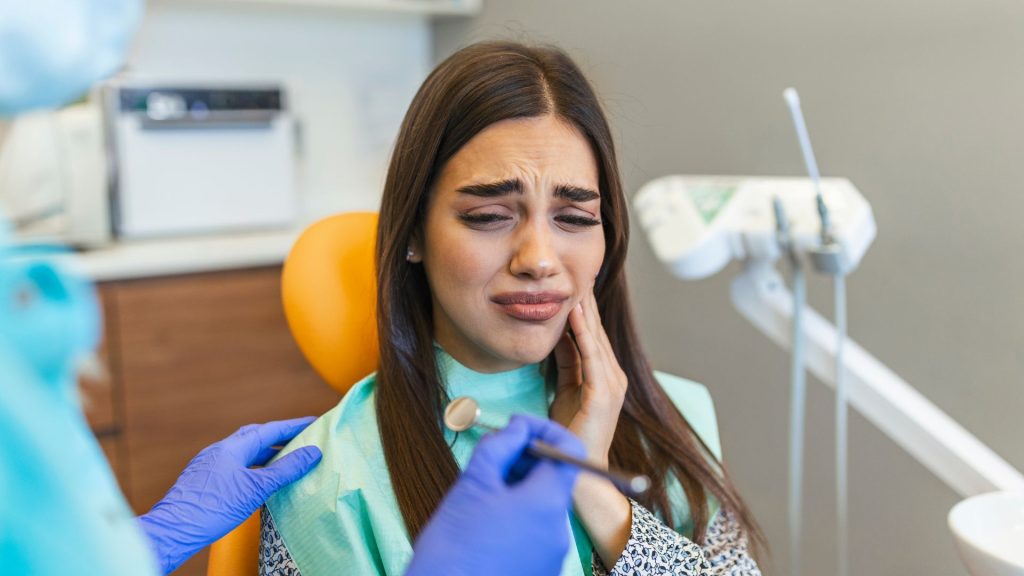 A woman sits in a dental chair touching her cheek with a pained expression while a dentist wearing gloves holds dental tools nearby.