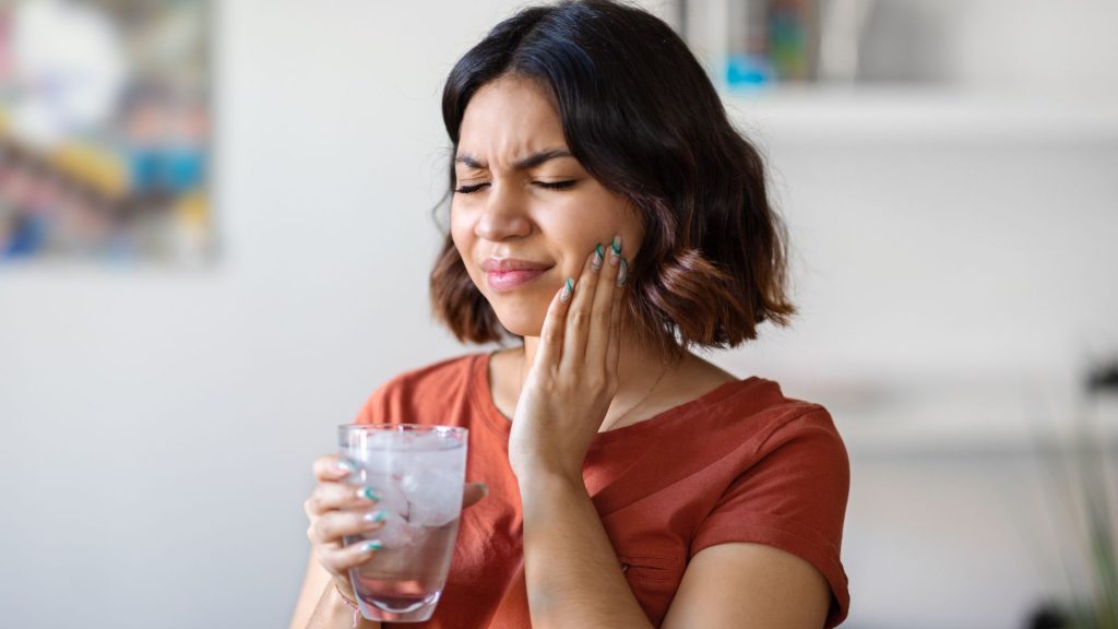 Woman holding a glass of ice water and touching her cheek, appearing to experience tooth pain or sensitivity.