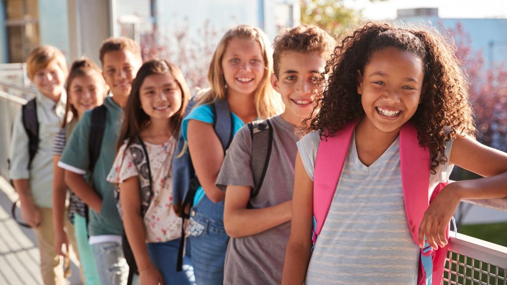 A group of school children with backpacks stand in a line outdoors, smiling at the camera.