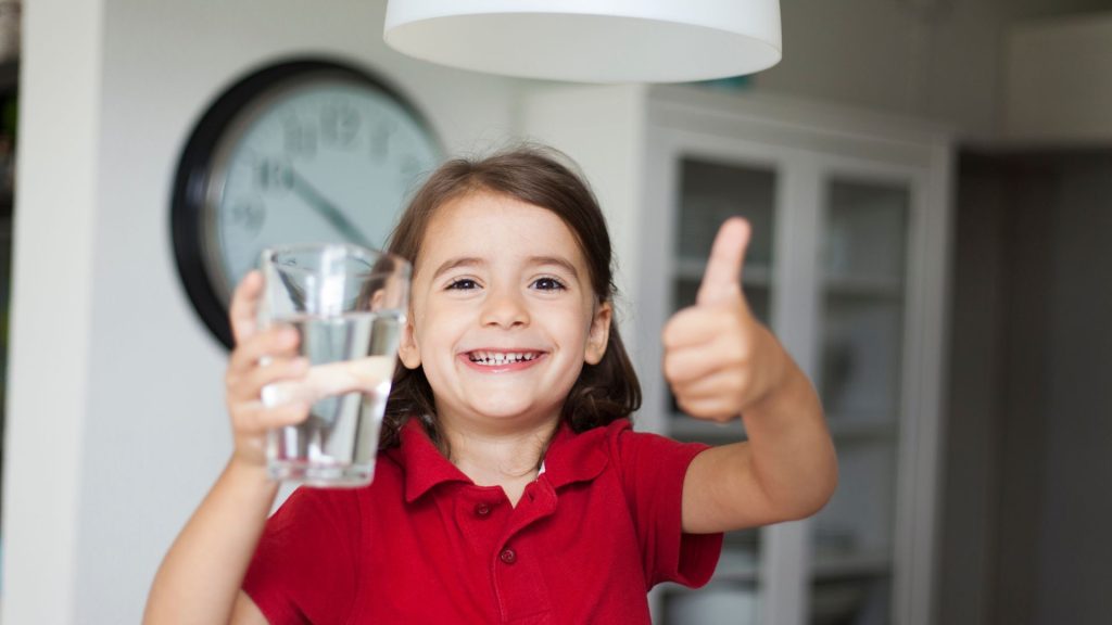 A child in a red shirt smiles, holding a glass of water in one hand and giving a thumbs-up with the other. A clock and cabinet are in the background.