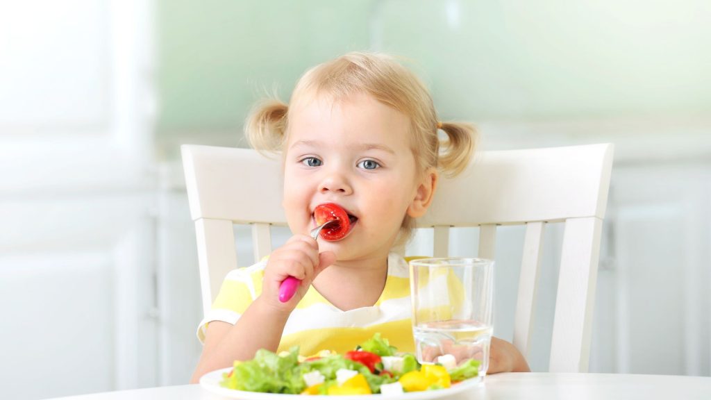 A young child with blond hair in pigtails sits at a table, holding a spoon near their mouth, with a plate of salad and a glass of water in front of them.