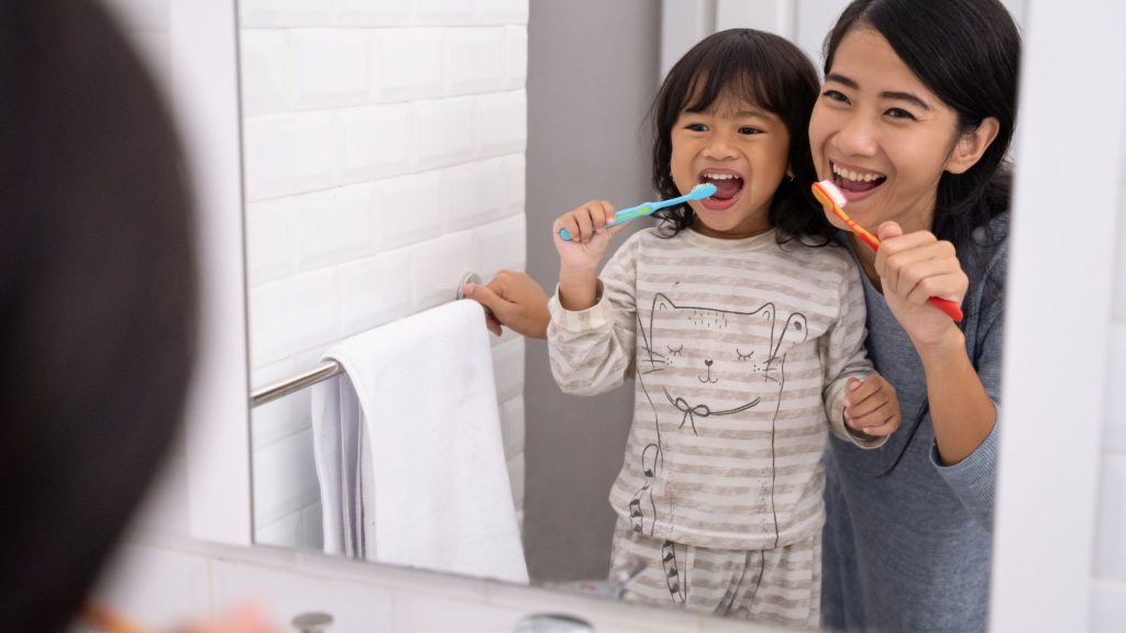 How to prevent cavities in kids - make dental care fun. An adult and a child are brushing their teeth together in front of a bathroom mirror. The child is wearing striped pajamas with a cat print.