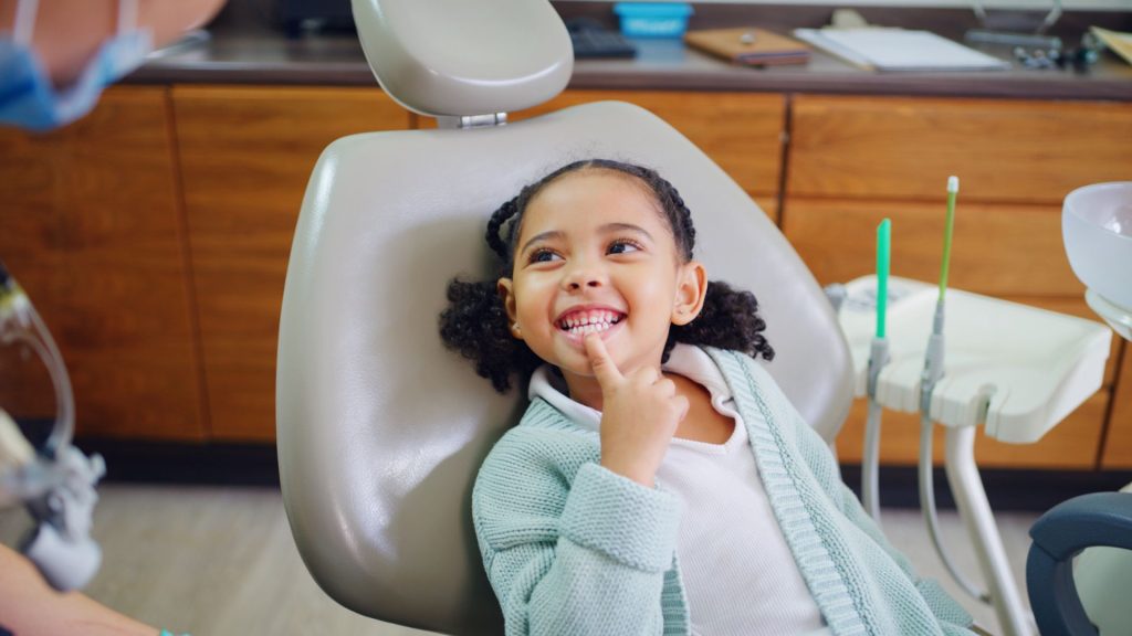A young girl smiles and points to her teeth while sitting in a dental chair at a dentist's office.