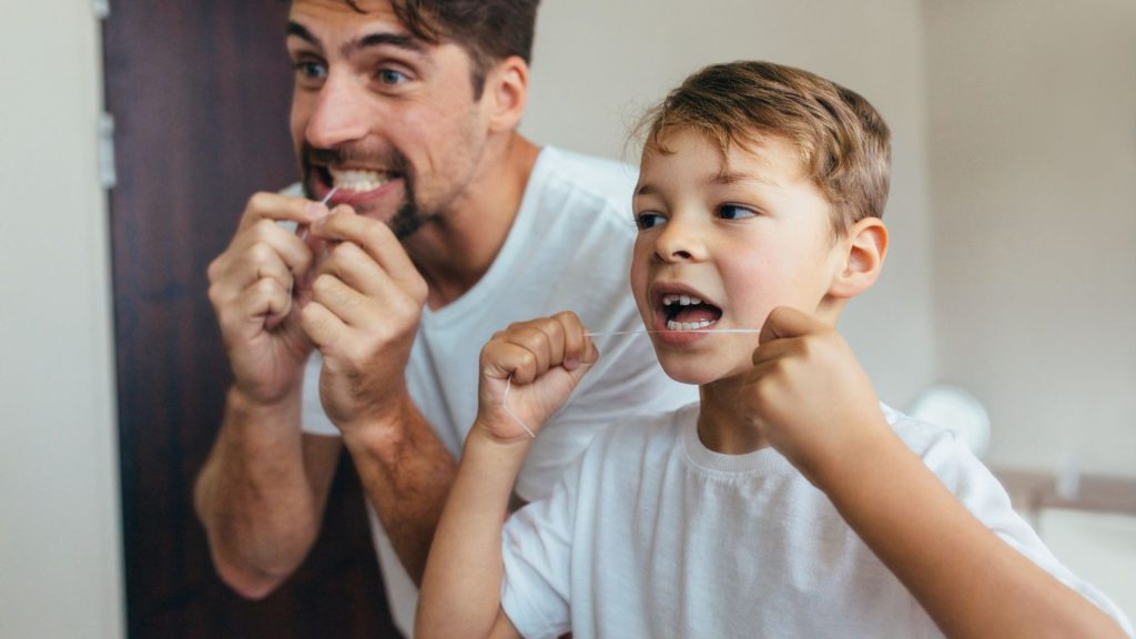 An adult and a child stand side by side in a bathroom, both wearing white shirts and flossing their teeth in front of a mirror.