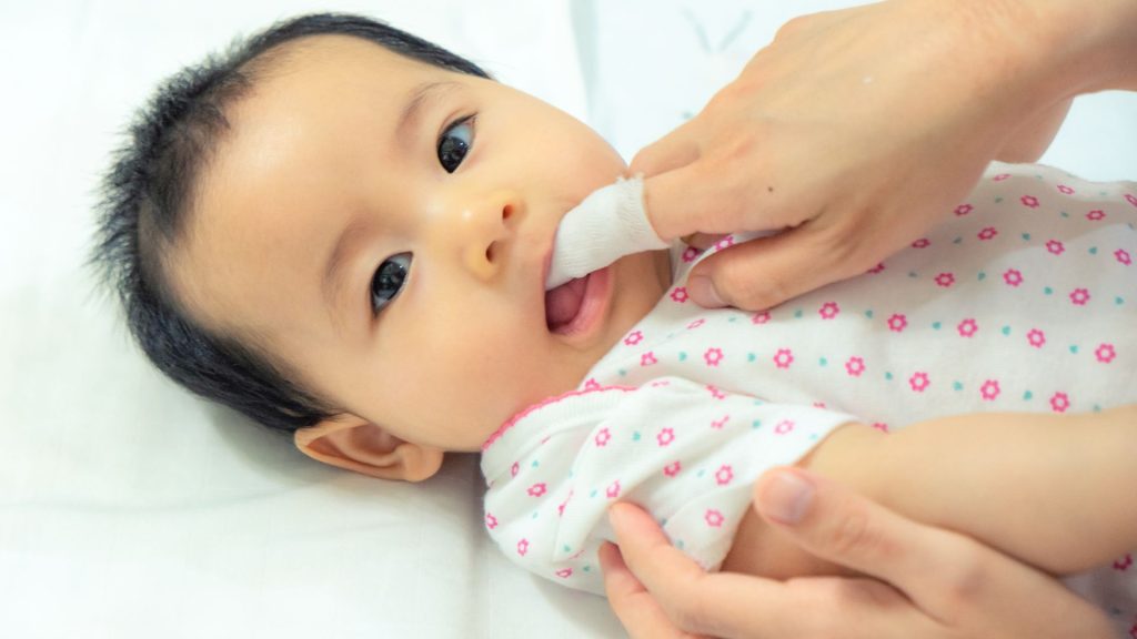 An infant lies on a white surface while an adult cleans the baby’s mouth with a soft cloth or gauze wrapped around their finger.