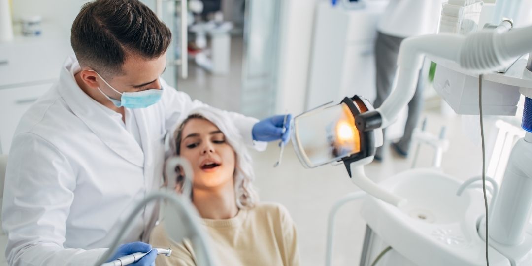 A dentist wearing a mask examines a woman's teeth in a dental clinic, using dental instruments and an overhead examination light.