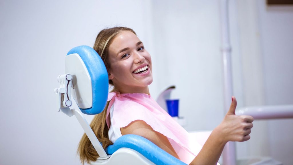Finding the right gentle dentist for you. A young woman sits in a dental chair, smiling and giving a thumbs up while wearing a pink dental bib in a clinic setting.