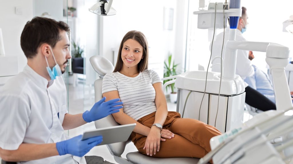 How a gentle approach improves your experience. A dentist wearing gloves and a mask consults with a smiling female patient seated in a dental chair in a modern clinic.