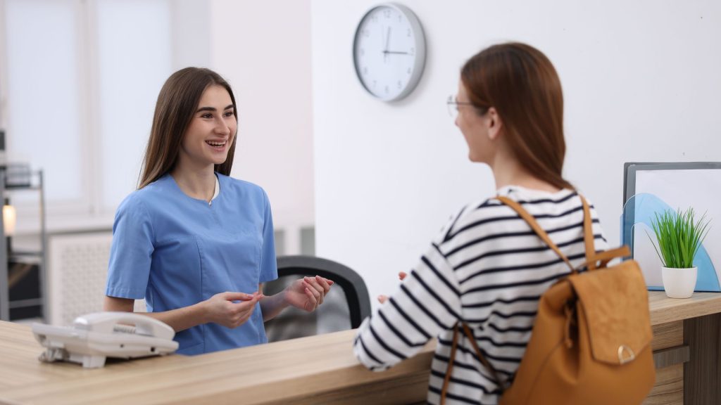 Tips for nervous patients before your appointment. A receptionist in blue scrubs smiles and talks to a woman with a backpack at a front desk. A clock, phone, and potted plant are visible in the background.