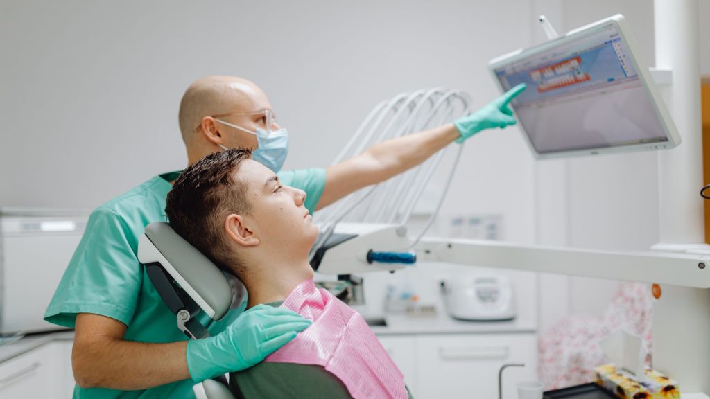 What makes a dentist gentle? A dentist wearing gloves and a mask points to a dental X-ray on a monitor while a young male patient in the chair looks at the screen.
