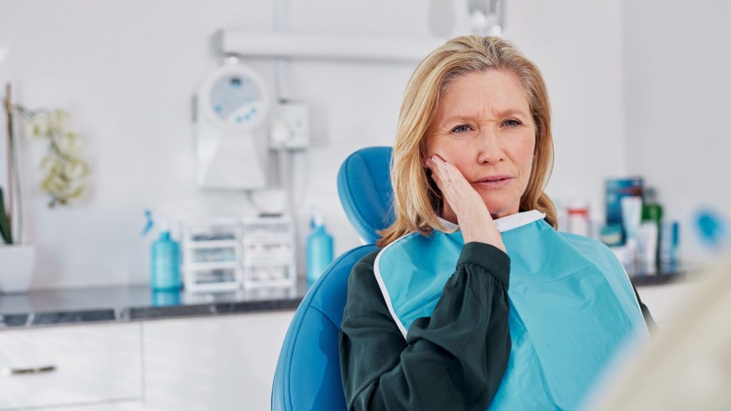 Is tooth sensitivity serious? When you should see a dentist for tooth sensitivity. A woman sits in a dental chair, touching her cheek and appearing concerned, in a modern dental clinic setting.