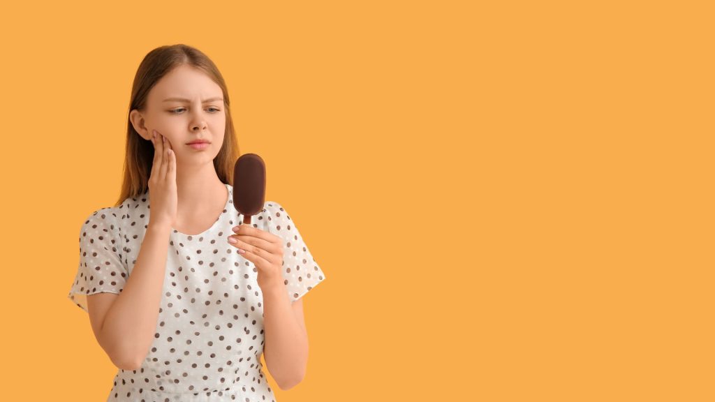 When tooth sensitivity is not serious. A young woman in a polka dot shirt holds her cheek and looks at a chocolate-covered ice cream bar with a concerned expression against an orange background.