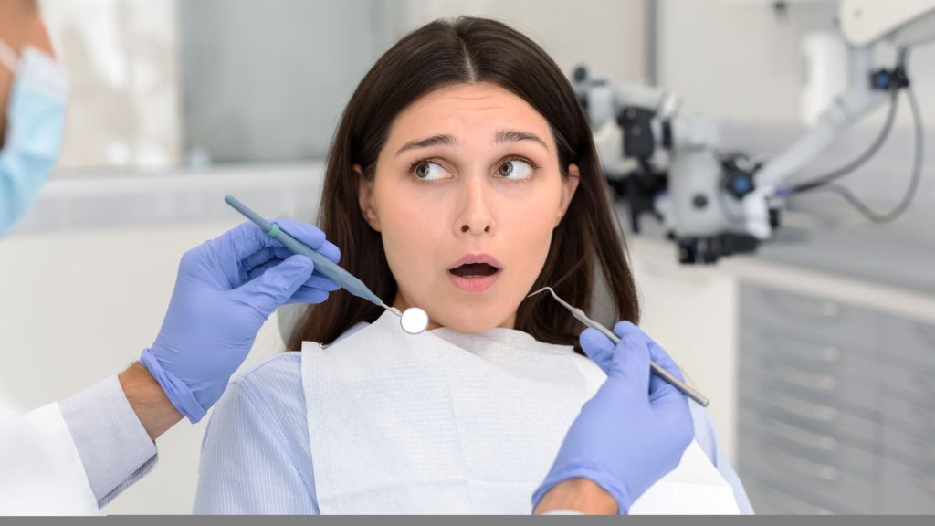 Why dental anxiety is more common than you think. A woman sits in a dental chair looking surprised or anxious while a dentist holds dental tools near her mouth.