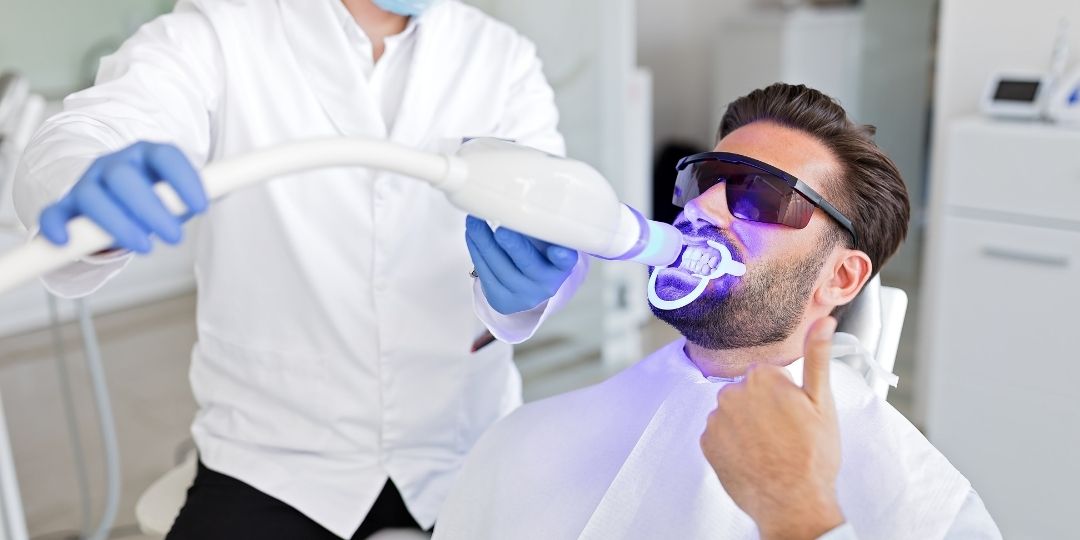 zoom whitening teeth whiteing A man wearing protective glasses receives a teeth whitening treatment with a UV light device at a dental clinic and gives a thumbs-up gesture.