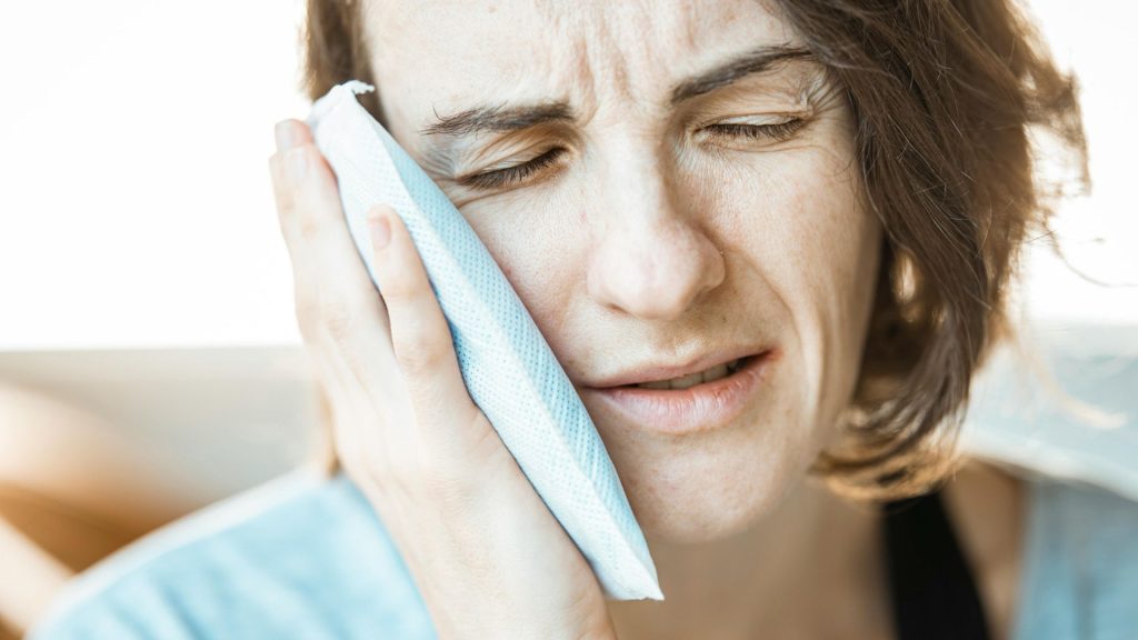 A person holding an ice pack to their cheek with a pained expression, suggesting jaw pain discomfort or pain in the facial area.