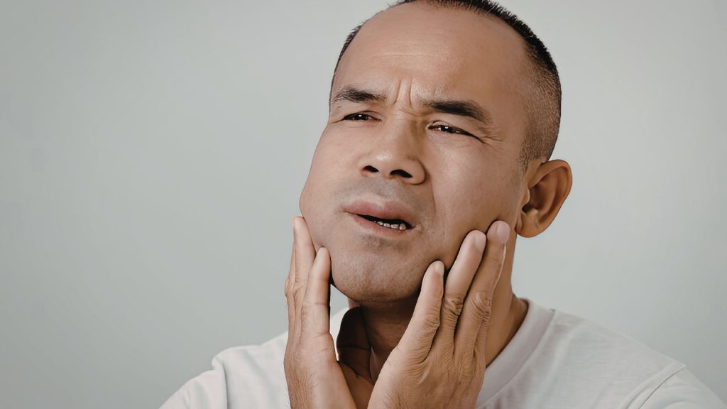 A man holds his jaw with both hands and appears to be in pain or discomfort, possibly indicating trismus or jaw pain.