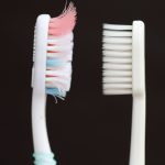 Two toothbrushes side by side: the left one has frayed, multicolored bristles, while the right one has straight, white bristles, both against a dark background.