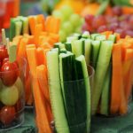 Clear plastic cups filled with cucumber sticks, carrot sticks, cherry tomatoes, and grapes are arranged on a green tablecloth. A plate of assorted fruit is in the background.